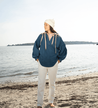 Person wearing a blue jacket and white pants standing on a beach with water in the background